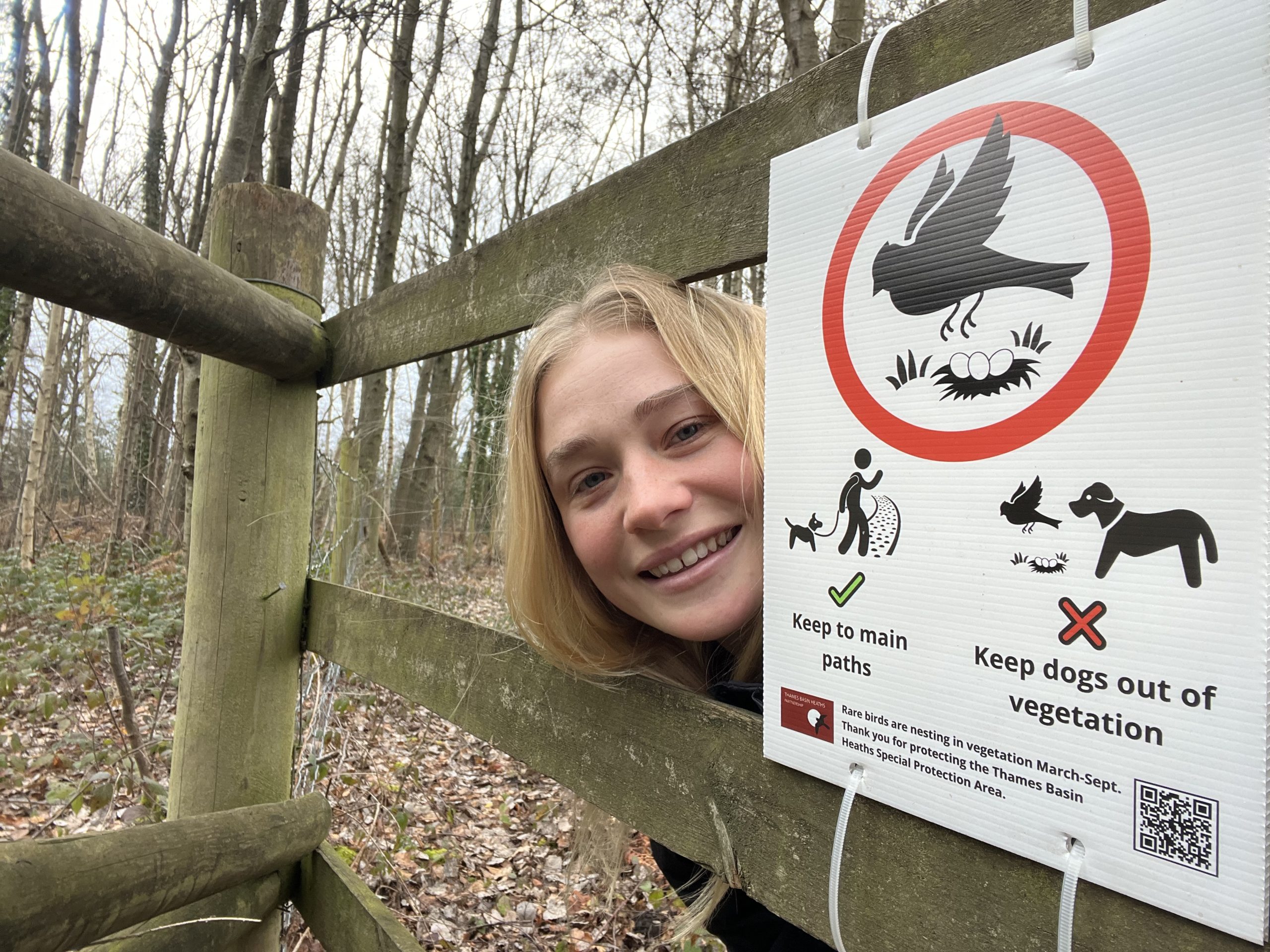 Photo of a girl with long blond hair pokes her head out from behind a sign that says "Keep to main paths".