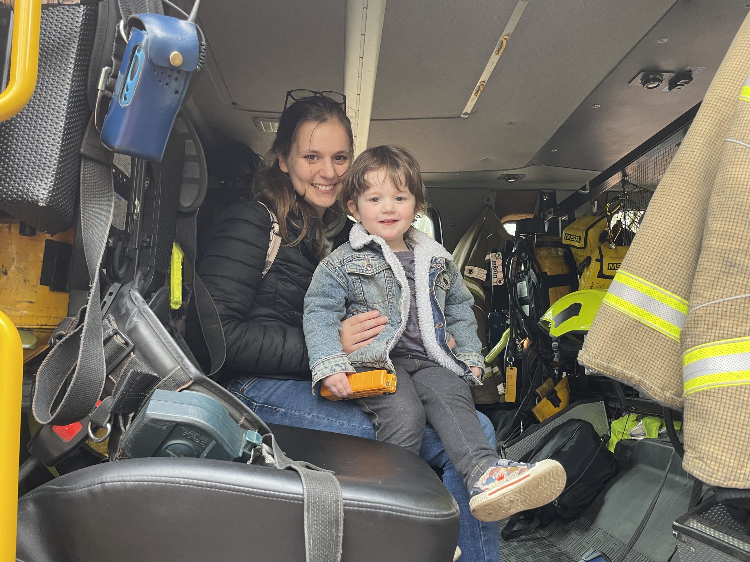 A photo of a small child sitting on his mum's lap, inside the cab of a fire engine. The child looks very happy.