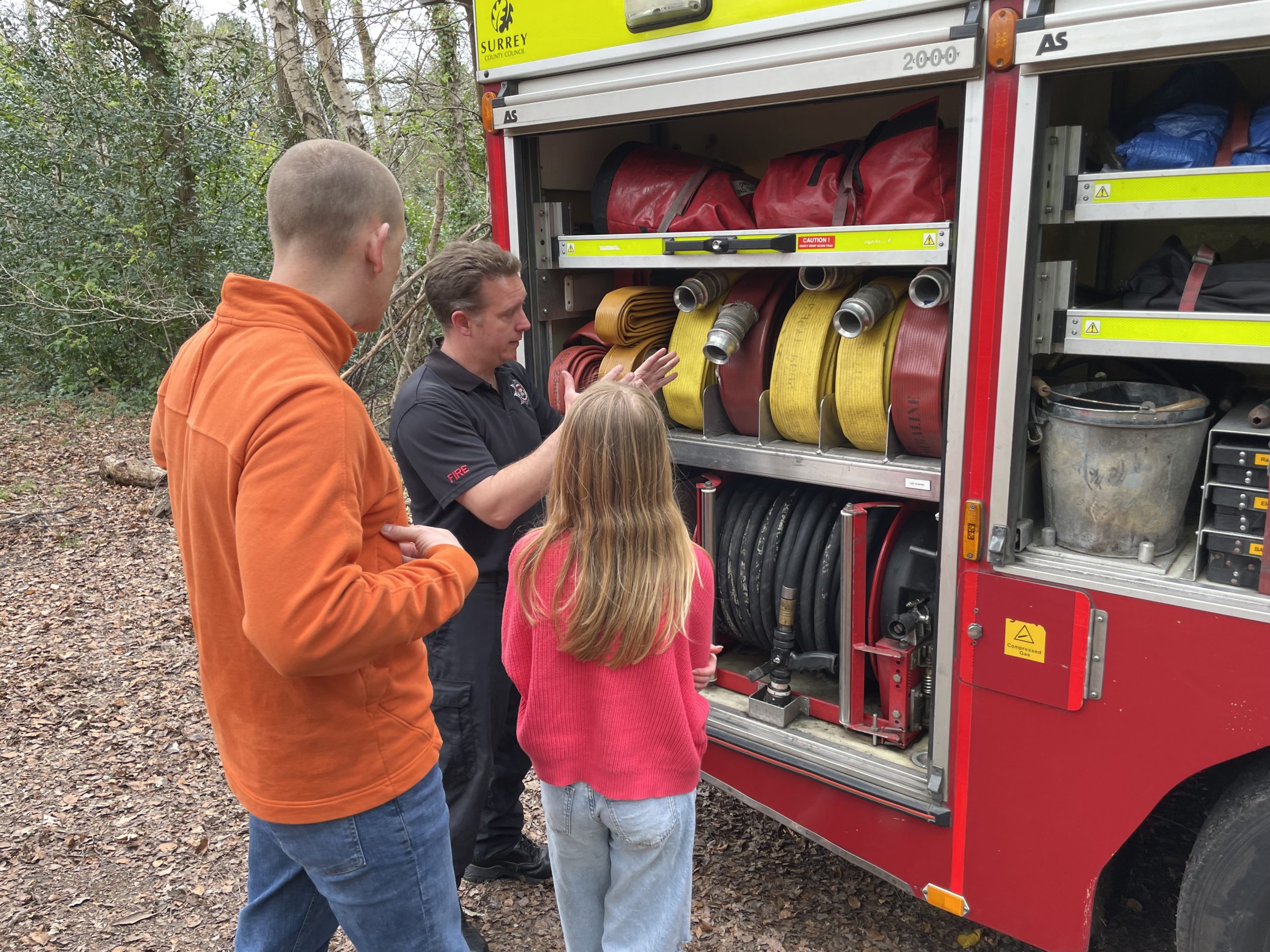Photo of a man in an orange top and a girl in a red top being shown the hoses on a fire engine.
