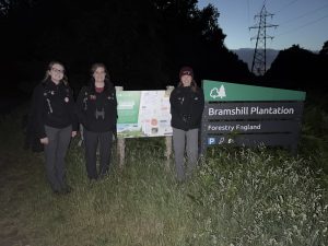 Photo of senior warden Kay and seasonal wardens sophie and sarah posing in from of a Bramshill Plantation sign in the dark. Taken after a Nightjar event.